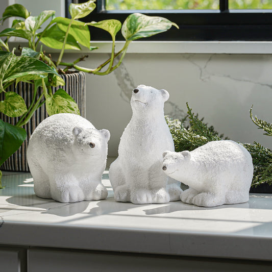 A set of three ceramic figurines of polar bears in different sizes with glittered details, placed on a surface next to a window with plants.
