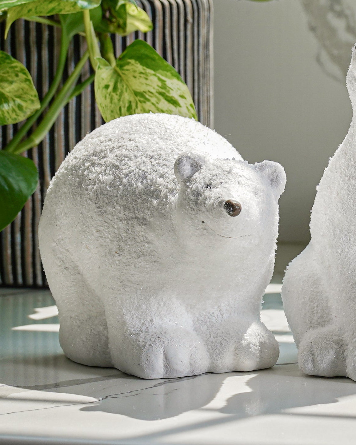 A set of three ceramic figurines of polar bears in different sizes with glittered details, placed on a surface next to a window with plants.