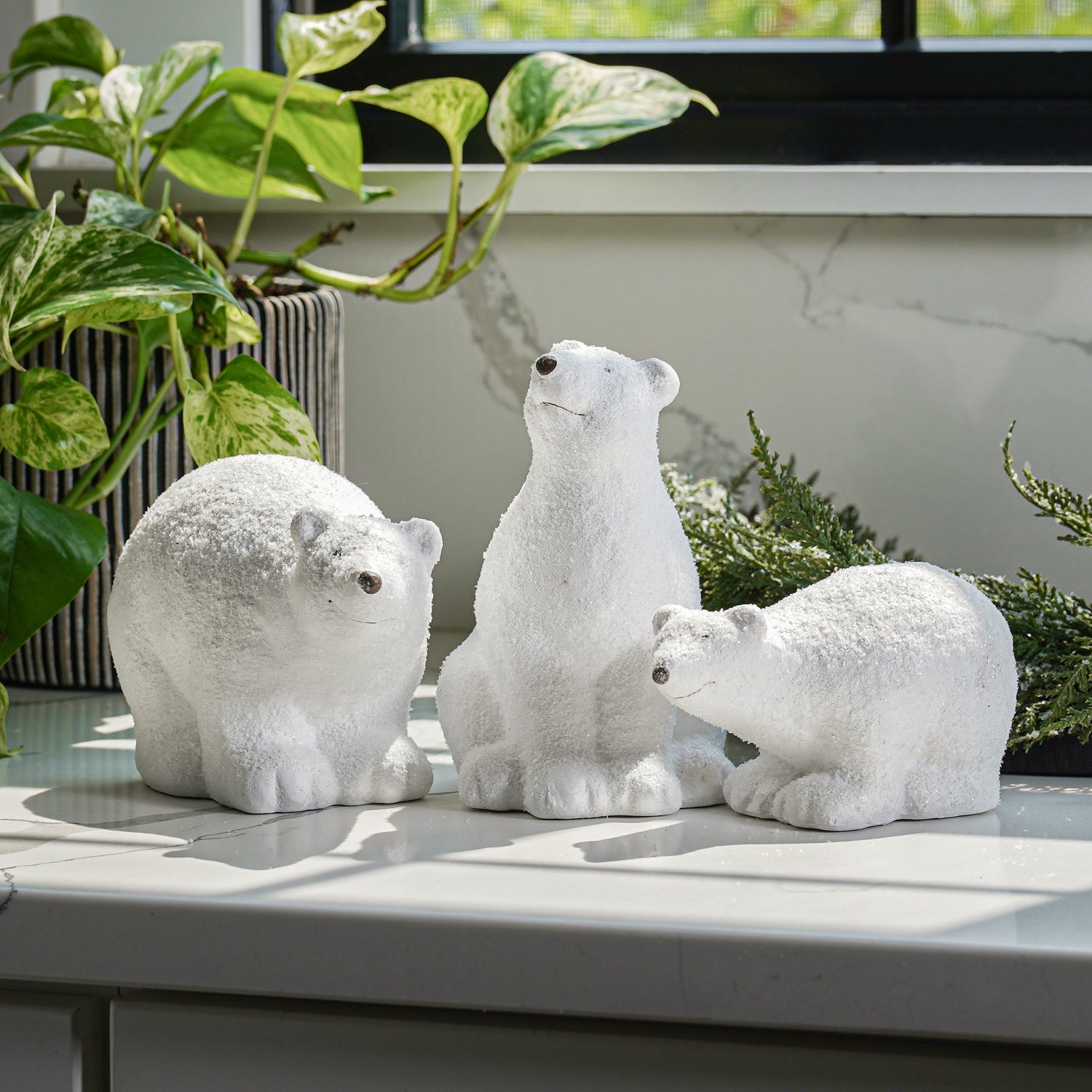 A set of three ceramic figurines of polar bears in different sizes with glittered details, placed on a surface next to a window with plants.