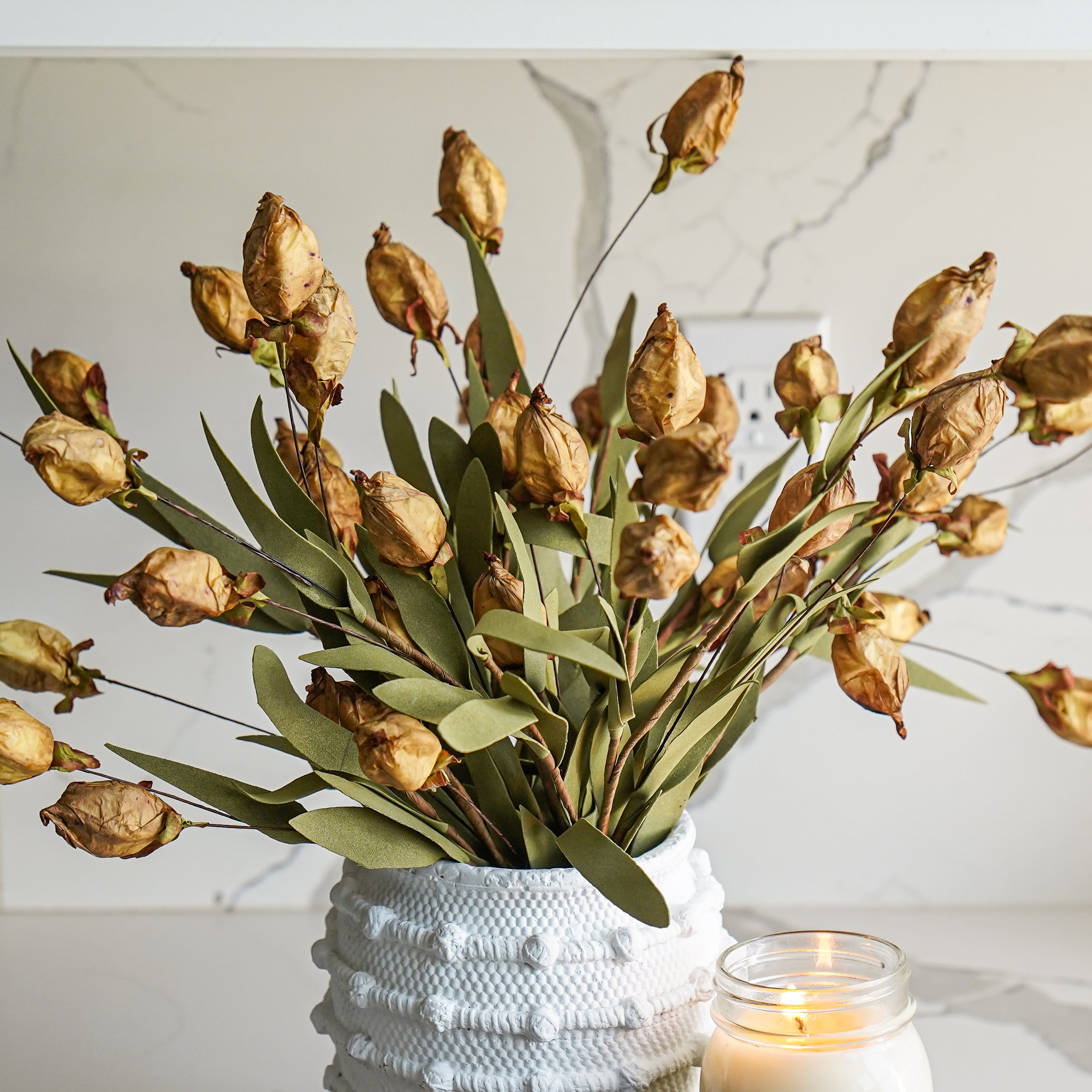 A faux buckskin color lantern bud bush with dried leaves in a white vase, placed on a marble surface with a candle beside it.