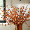 A burnt orange faux leafy berry bush with a collection of vibrant orange leaves and dark orange berries, arranged in a glass vase, placed next to a window.