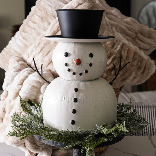 A painted tin snowman with a black top hat and a scarf, standing on a round metal base, decorated with a green wreath and white artificial snow.