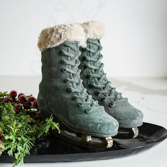 A pair of beige velvet ice skates with faux fur trim, displayed on a black stand with greenery and resin details.