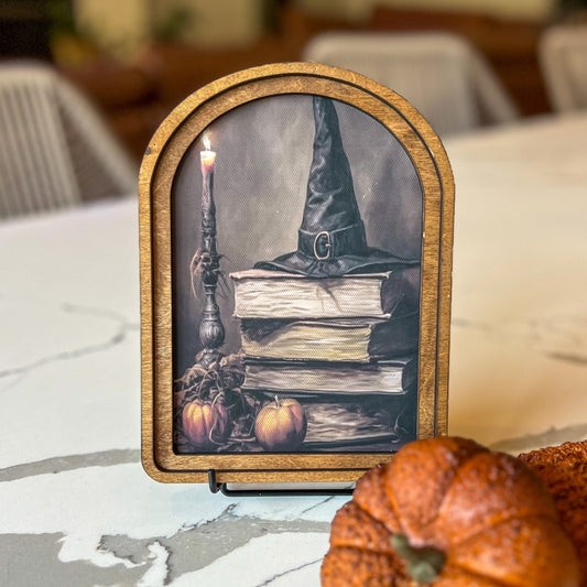 Decorative sign with a witch hat, books, and pumpkins on a table.