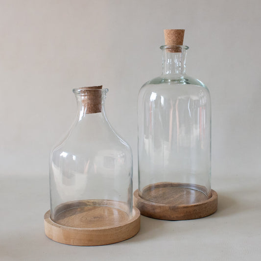 Two clear glass bottles with wooden bases and cork lids on a light gray background