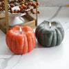 Two pumpkin-shaped candles, one orange and one green, on a marble surface with a decorative branch in the background.