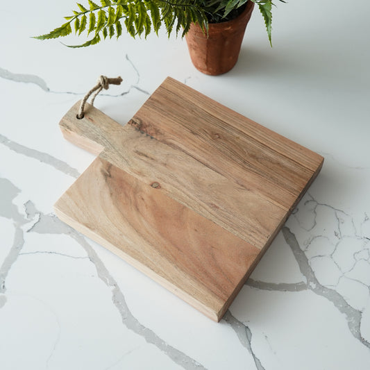 Wooden cutting board with a handle on a marble surface with a plant in the background