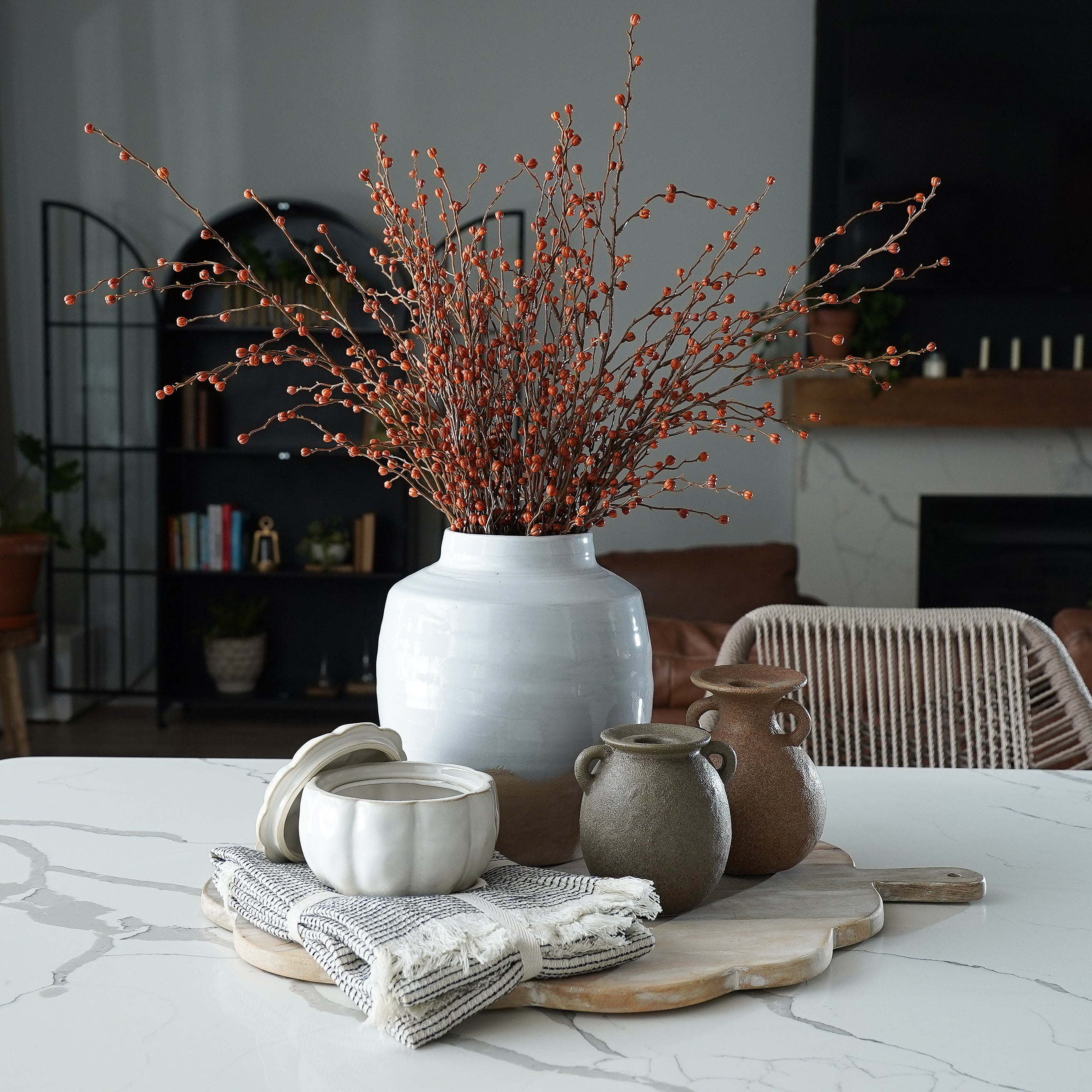 Decorative setting with a vase of dried branches, ceramic pots, and a wooden cutting board on a marble surface.
