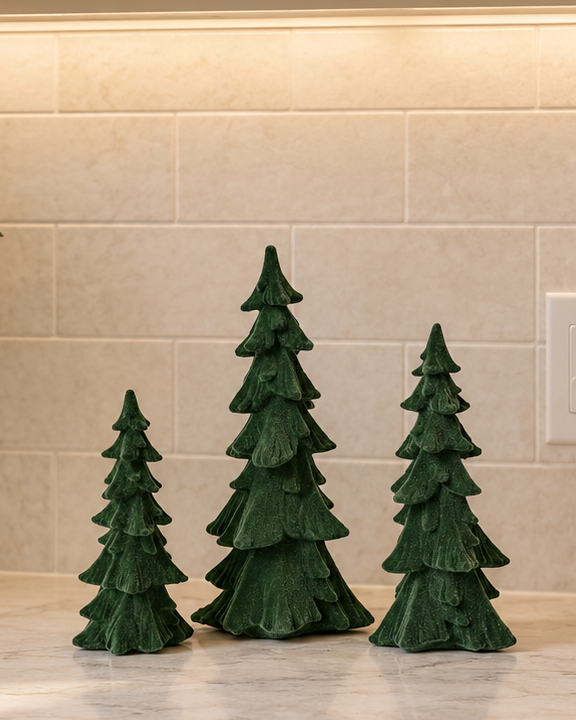 Set of three evergreen green resin Christmas trees with textured layered branches in small, medium and large sizes displayed on a kitchen countertop