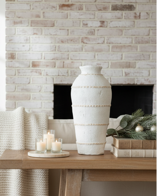 Decorative setting with a vase, candles, and a blanket on a wooden table against a brick wall.