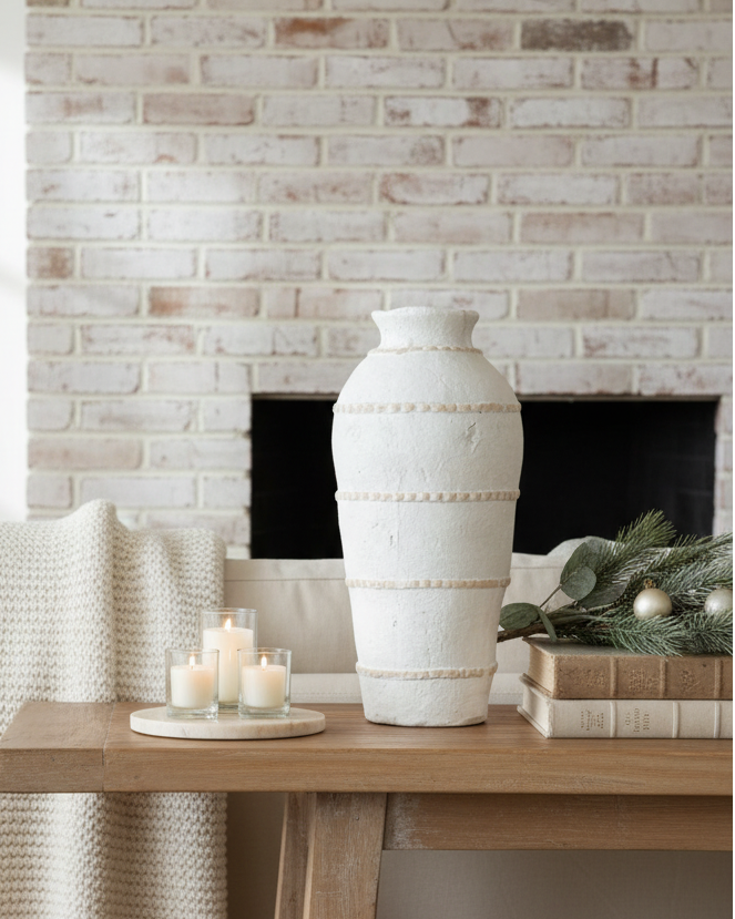 Decorative setting with a vase, candles, and a blanket on a wooden table against a brick wall.