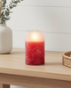 Candle with a red and white design on a wooden surface next to a white vase and a woven basket.
