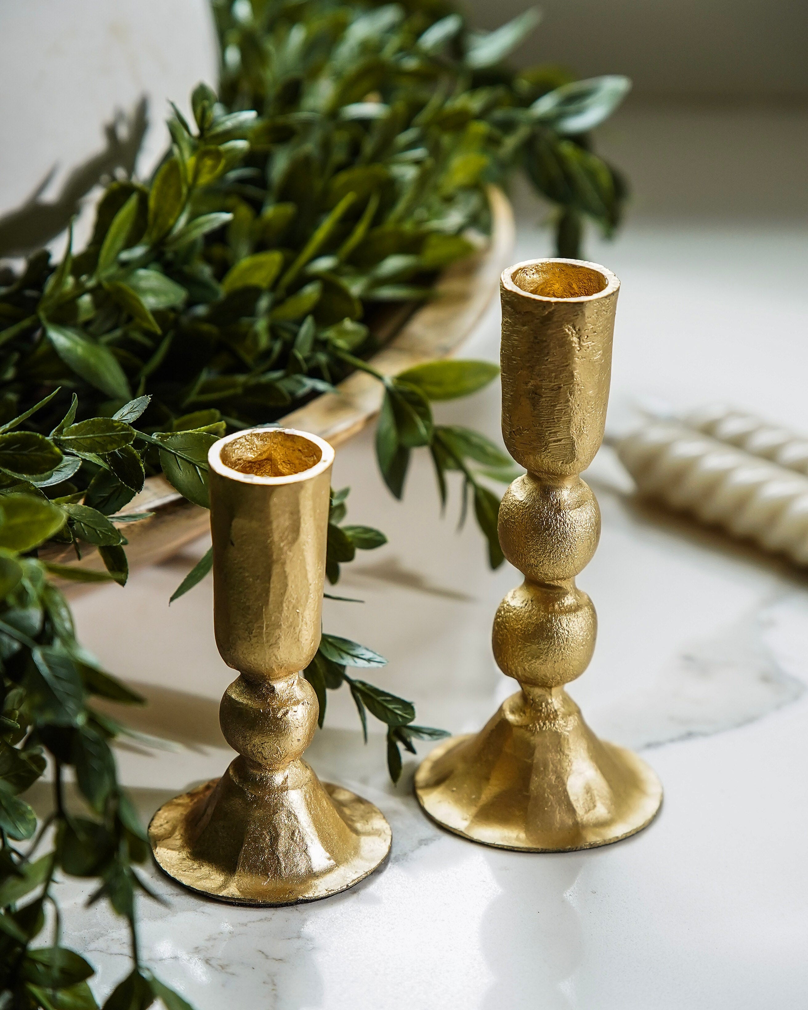 Two hammered gold candlesticks on a white marble surface, surrounded by green foliage and a white rope accent.