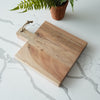 Wooden cutting board with a handle on a marble surface with a plant in the background