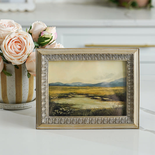 Framed landscape painting on a table with pink roses in the background