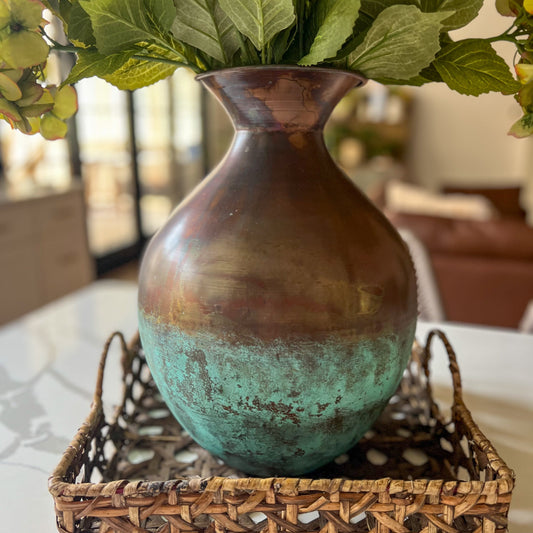 Bronze vase with greenish-blue patina on a woven tray with flowers in the background