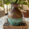 Bronze vase with greenish-blue patina on a woven tray with flowers in the background