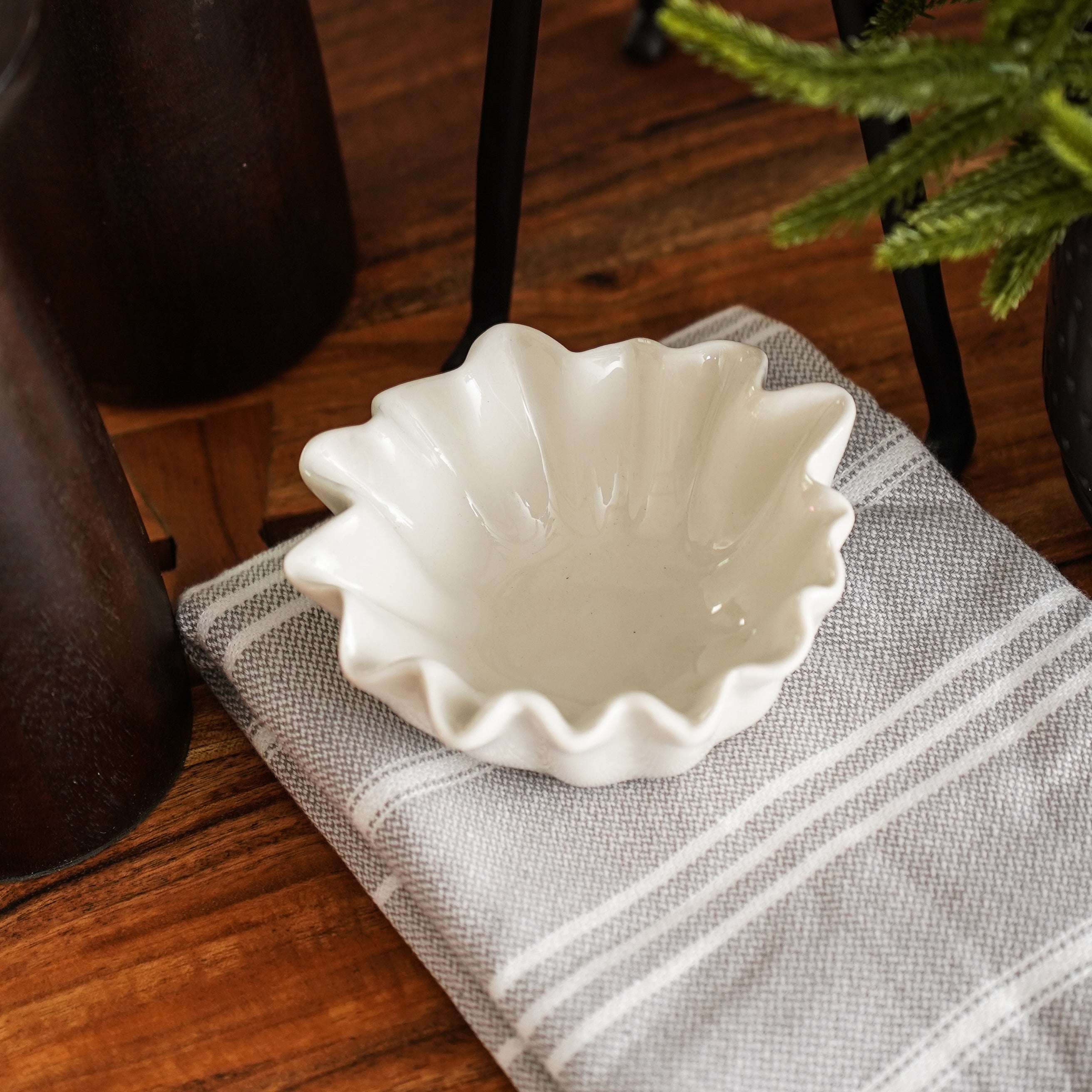 A white stoneware fluted bowl is displayed on a striped napkin, placed on a wooden surface, with a decorative plant in the background.